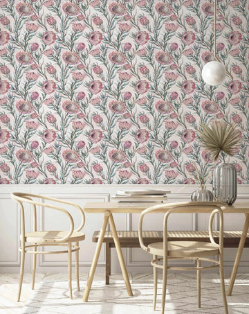 Protea on Blush Wallpaper displayed on a dining room wall above white wainscoting. The wallpaper features a repeating botanical pattern of pink and mauve protea flowers with green stems on a cream background, rendered in a detailed watercolor illustration style. Two natural wood chairs with woven seats are positioned at a wooden table beneath the wallpaper. A white pendant light hangs above, and a clear glass vase with dried grasses sits on the table. The overall aesthetic is soft, botanical, and classic.