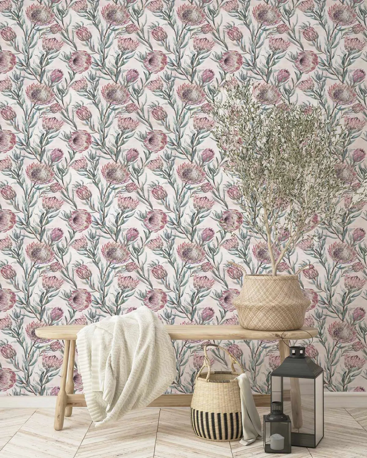 Protea on Blush Wallpaper displayed on a bedroom wall featuring a repeating botanical pattern of mauve and dusty rose protea flowers with green stems and foliage on a cream background. A light wood bench sits against the wallpapered wall with a cream throw blanket draped over it, a woven basket and striped decorative basket on the floor, and a black metal lantern with candle nearby. The wallpaper has a hand-drawn, watercolor illustration style with organic botanical details throughout.