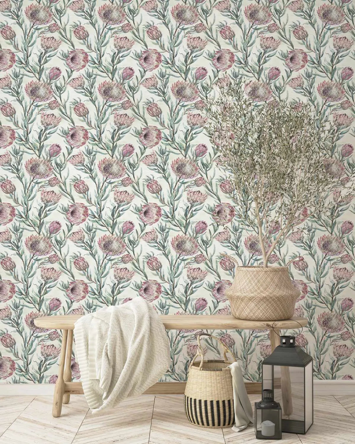 Wallpaper sample featuring a repeating botanical pattern of protea flowers in dusty mauve and soft pink tones with sage green stems and foliage on a cream background. The design is displayed on a wall behind a natural wood bench styled with a cream throw blanket, a woven basket containing dried pampas grass, a striped woven container, and a black metal lantern with a candle, creating a serene, botanical-inspired interior setting on light wood flooring.