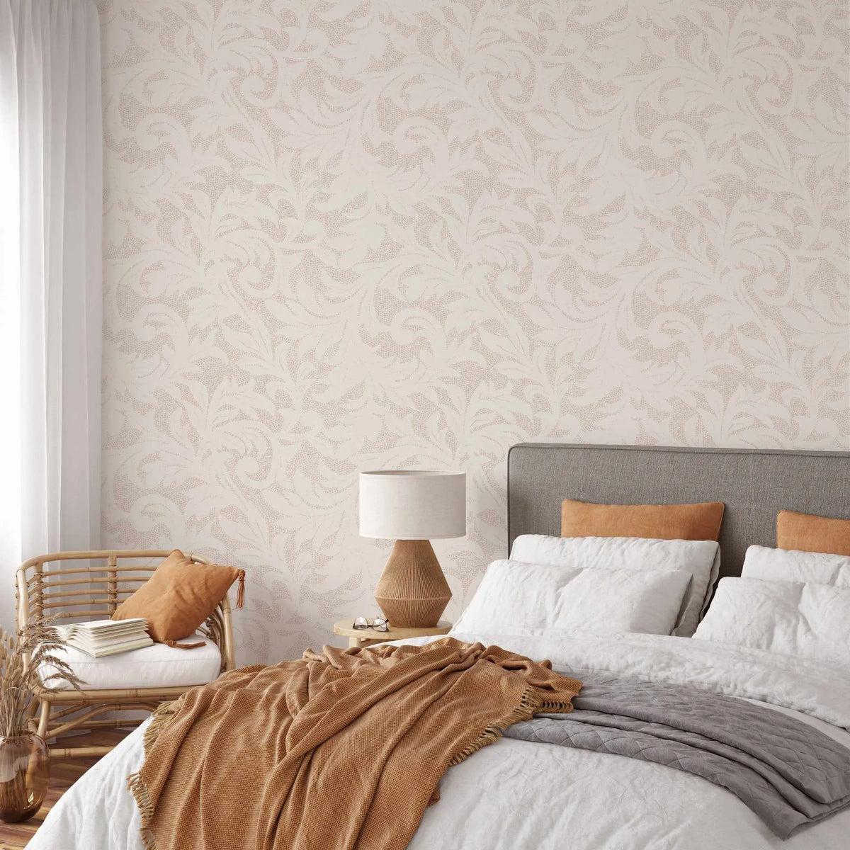 Provence in Sand Wallpaper displayed on a bedroom wall behind a gray upholstered bed. The wallpaper features a repeating classical damask pattern with ornate scrolls and leaf motifs in warm sand and cream tones on a light ivory background. The design has a subtle woven texture appearance. The bedroom setting shows the wallpaper paired with natural wood furnishings, tan and white bedding, a woven rattan chair with caramel cushions, and a white linen shade table lamp, creating a warm, neutral aesthetic.