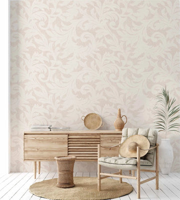 Lifestyle shot of a living room featuring Provence in Sand wallpaper. The wallpaper has a repeating pattern of organic, swirling leaves and vines in a subtle, light beige tone against an off-white background, with a textured, dotted effect within the leaf shapes. The room includes a light wood sideboard with books, a woven basket, a round jute rug, a light wood chair with a straw hat, a large white vase with a green plant, and a decorative ceramic jug.