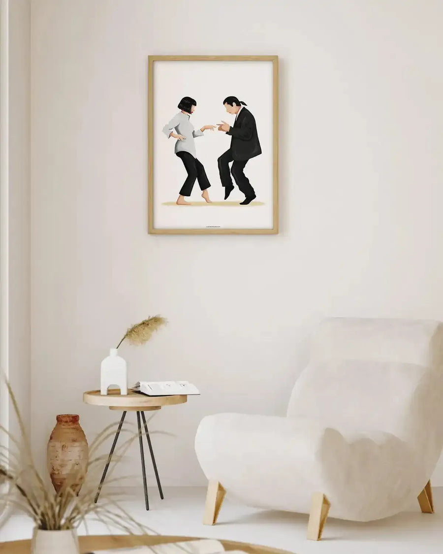 Large framed art print displayed above a small wooden side table in a minimalist living room. The print features an illustration of two figures, a woman with a black bob and a man with dark hair tied back, dancing in black and white outfits on a light background, with a natural wood frame and white border. The room includes a cream armchair, a terracotta vase, and dried pampas grass in a white ceramic vase.