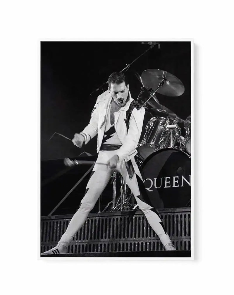 Black and white framed canvas art print featuring a performer in a white suit and black shirt mid-performance, captured in an energetic pose with one leg extended. A drum kit is visible in the background with "QUEEN" text on the bass drum. The photograph has high contrast with deep blacks and bright whites, mounted in a white frame with white border, presented as a portrait-orientation wall art piece.