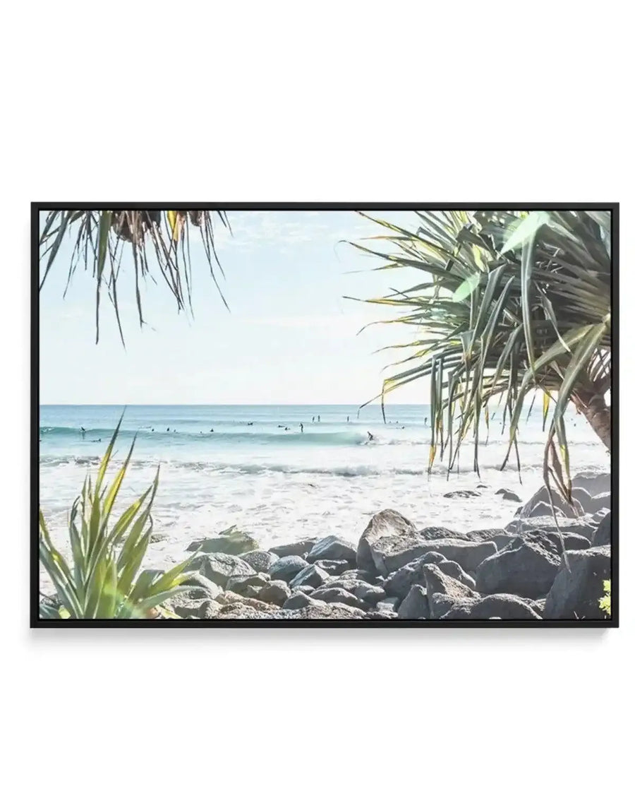 Framed canvas print of a vibrant coastal beach scene with a black frame. The composition features a light blue sky with scattered clouds, a turquoise ocean with gentle white waves, and numerous surfers scattered across the water. The foreground shows a rocky shoreline with light-coloured sand and green tropical plants on either side, framing the view of the ocean.