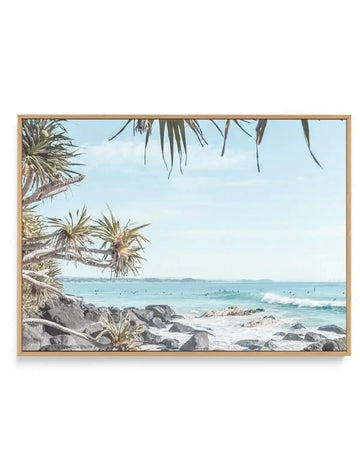 Framed canvas art print of a coastal beach scene with a light natural wood frame. The photograph features a light blue sky with wispy clouds, a turquoise ocean with gentle waves and surfers, and dark grey rocks on the shore with some green foliage.
