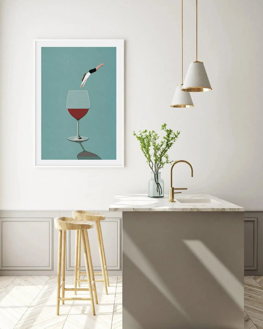 Kitchen:
Framed art print displayed on a white wall above a kitchen island with two wooden bar stools. The print features a minimalist illustration of a person diving headfirst into a glass of red wine, set against a teal background. The kitchen has white walls with wainscoting, a light-colored marble countertop, a gold faucet, and two pendant lights with concrete shades and gold accents.