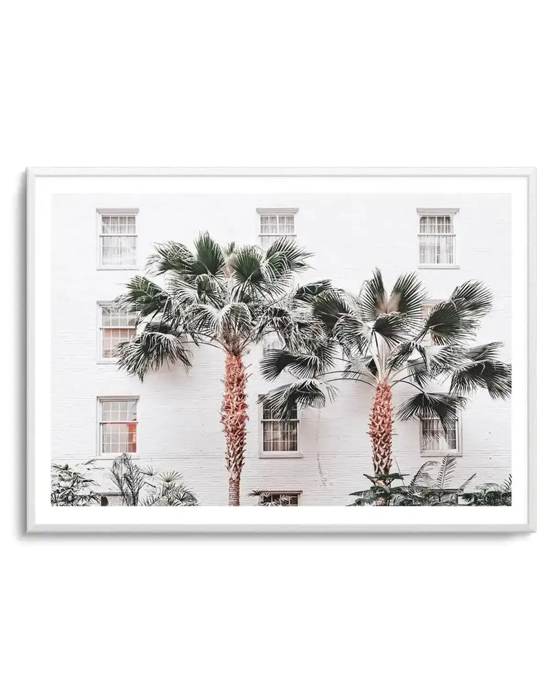 Framed photograph of two palm trees with vibrant green fronds and textured reddish-brown trunks, set against a white brick building with multiple windows. The composition captures a bright, tropical mood with a minimalist aesthetic.