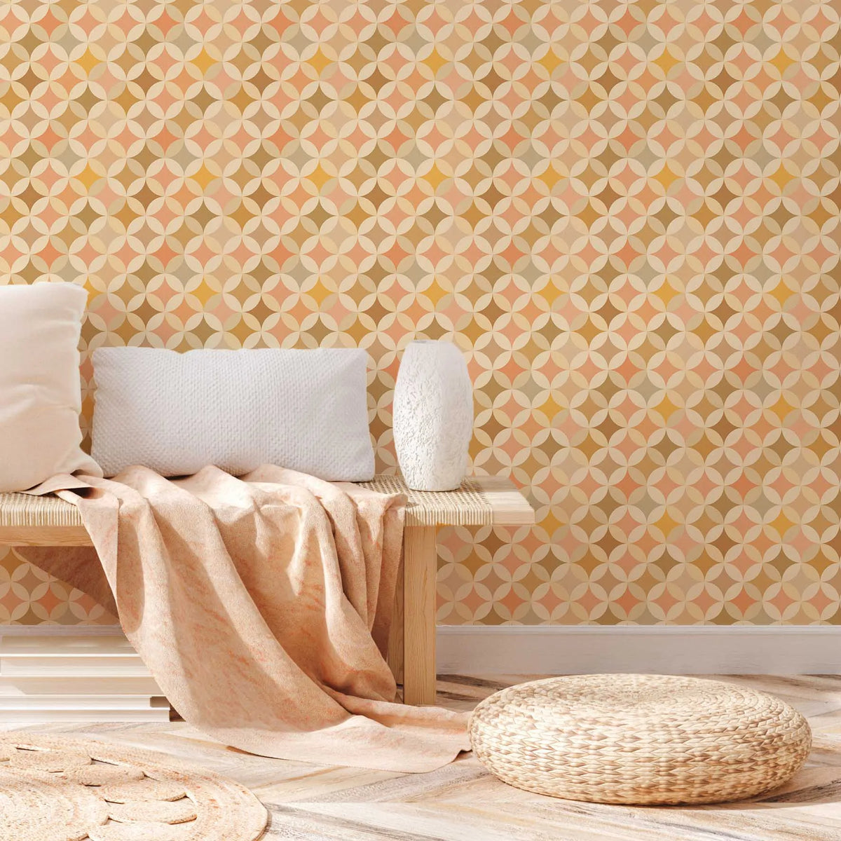 Retro Diamonds Wallpaper displayed on a bedroom wall behind a wooden bench. The wallpaper features a repeating geometric pattern of four-petaled diamond shapes in warm tones including cream, peach, coral, golden yellow, and sage green on a soft beige background. A white textured pillow and rolled white ceramic object sit on the bench with a peach linen throw draped over it. A woven round floor cushion and striped area rug are visible in the foreground, creating a cohesive warm, neutral aesthetic.