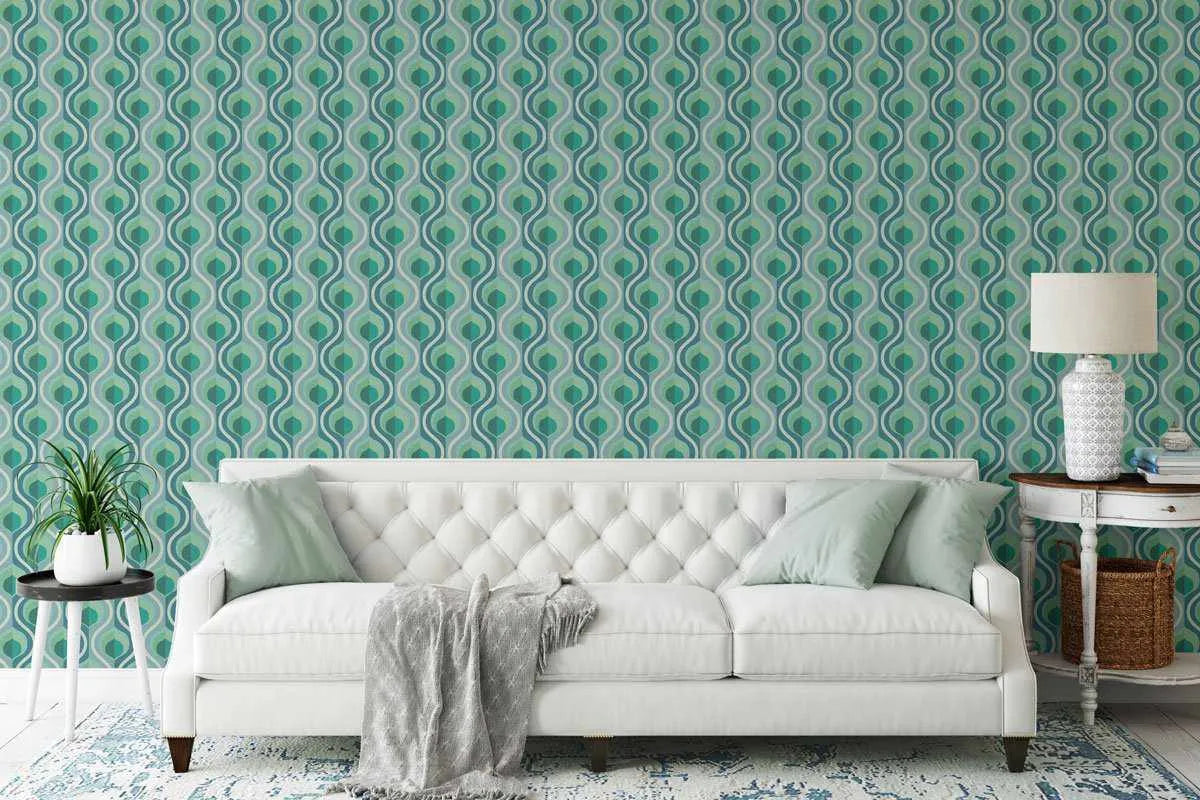 Retro Fun Wallpaper displayed on a living room wall behind a cream-colored tufted sofa. The wallpaper features a geometric repeating pattern of vertical teardrop or leaf shapes in shades of teal, seafoam green, and cream, creating an interlocking ogee design typical of mid-century modern style. The pattern has a subtle texture and matte finish. The room includes light wood side tables, a white ceramic table lamp, potted plant, and a patterned area rug in complementary teal and cream tones.