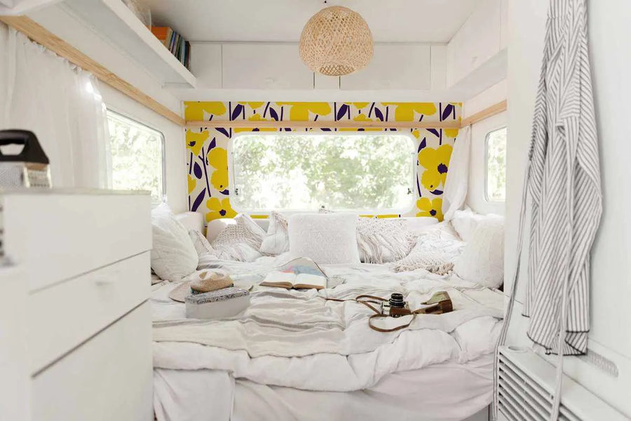 Retro Poppies Wallpaper displayed in a camper interior with bright yellow background and bold navy blue poppy flowers with dark centers. The wallpaper frames a large window with natural greenery visible outside. The design features a mid-century modern aesthetic with stylized floral motifs in a repeating pattern. The wallpaper is installed above the bed area in the compact living space, creating a cheerful focal point against white cabinetry and neutral bedding.