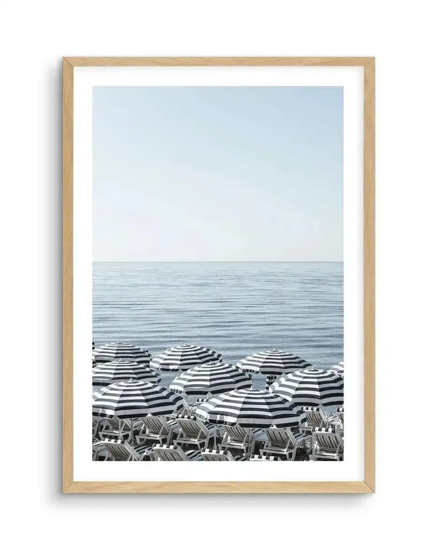 Framed photography art print of a beach scene with natural wood frame and white border. The composition features rows of black and white striped parasols and matching lounge chairs on a pebbled beach, with calm blue ocean and clear pale blue sky stretching to the horizon.