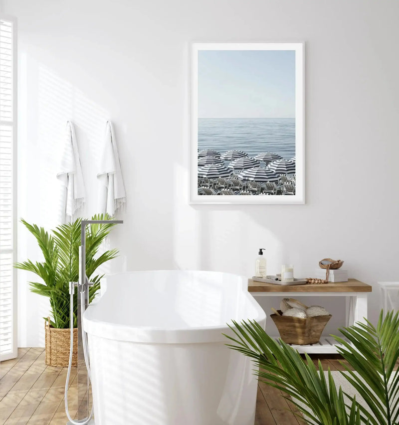 Lifestyle shot of a bright white bathroom with a large framed art print of a beach scene displayed above a white freestanding bathtub. The print features a calm blue ocean under a pale sky, with rows of black and white striped parasols and beach chairs on the sand, framed in white with a white border. The bathroom has light wood flooring, a large green plant in a woven basket, white towels hanging on the wall, and a wooden side table with bath accessories.