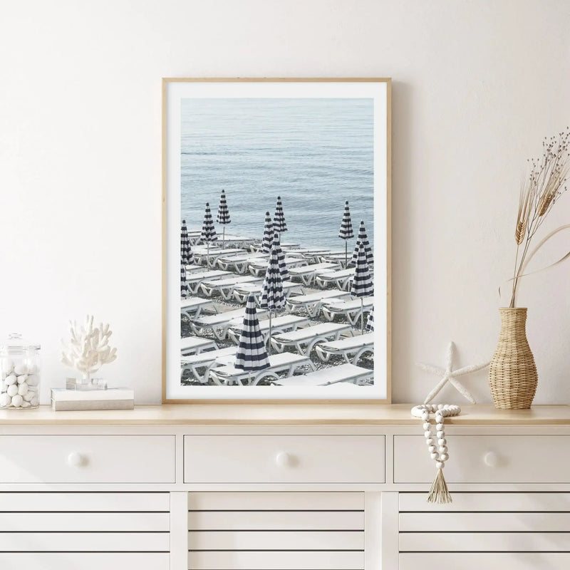 Framed art print displayed on a white dresser showing a beach scene with rows of striped navy and white parasols and matching lounge chairs arranged on sand, with calm ocean and sky in soft blue tones. Natural wood frame with white border. Styled with coastal décor including white coral, woven vase with dried grasses, pearl beaded garland, and white decorative objects.