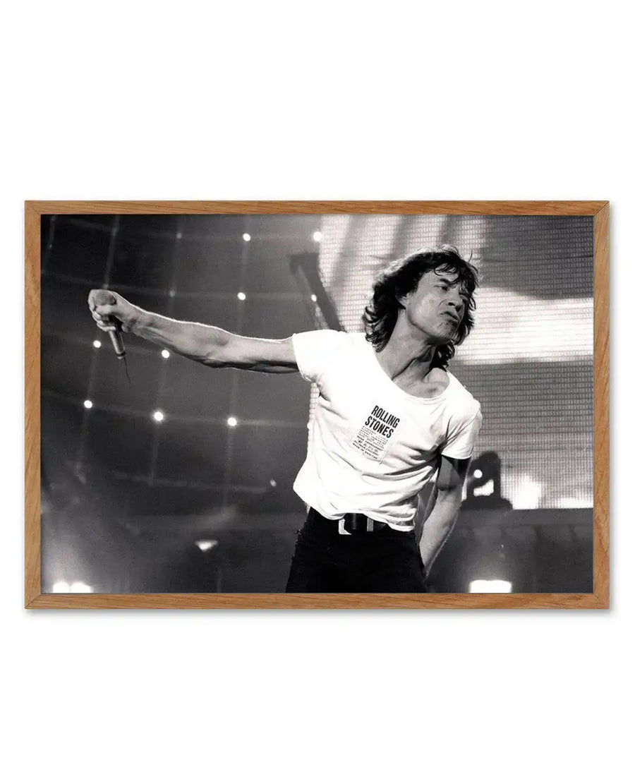 Framed black and white photograph of Mick Jagger performing on stage, captured mid-song with his arm extended, holding a microphone. He is wearing a white t-shirt with "ROLLING STONES" visible on the chest, and black pants. The background features stage lights and a large screen with a grid pattern, all displayed in a natural oak frame with a white border.