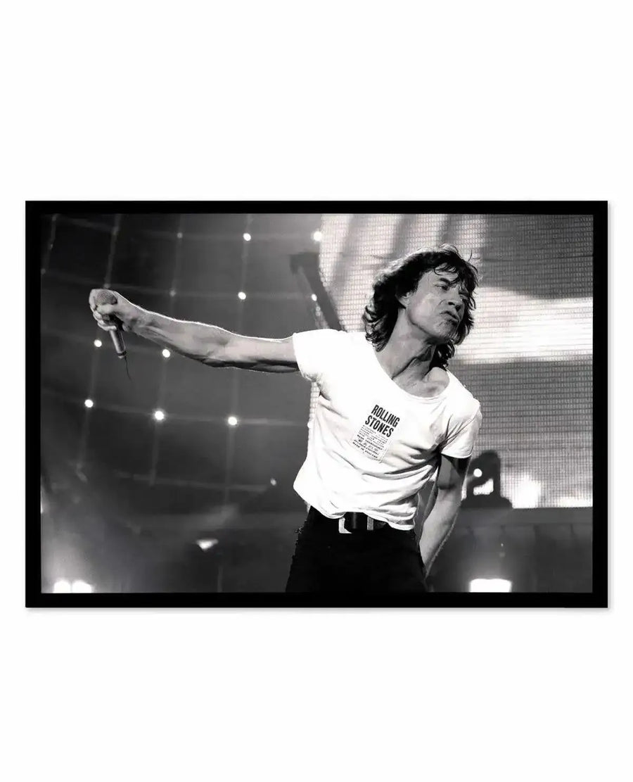 Framed black and white photograph of Mick Jagger performing on stage, captured in a dynamic pose with his arm extended, holding a microphone. He is wearing a white t-shirt with "ROLLING STONES" printed on it and dark trousers, with a black frame and white border. The background features stage lights and a large screen.