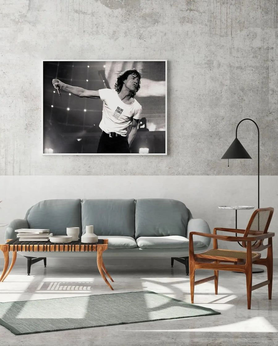 Lifestyle shot of a minimalist living room featuring a framed black and white art print of Mick Jagger performing live, displayed above a light grey sofa. The print captures Jagger mid-performance, holding a microphone, with his arm outstretched and wearing a white t-shirt with "ROLLING STONES" text, against a backdrop of stage lights. The room includes a light wood coffee table with books and ceramic vases, a dark wood and cane armchair, a black floor lamp, and a light grey rug on a concrete floor.