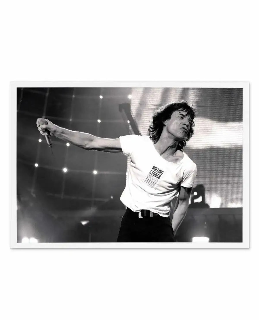 Black and white photograph of Mick Jagger performing on stage, captured mid-song with his arm extended holding a microphone. He wears a white t-shirt with "ROLLING STONES" printed on it, and black pants. The background features stage lights and a large screen, creating a dynamic concert atmosphere.