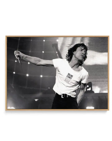 Framed black and white canvas print of Mick Jagger performing live on stage, captured mid-song with a microphone in his outstretched left hand. He wears a white t-shirt with "ROLLING STONES" printed on it, black trousers, and a black belt. His long hair is tousled, and his face shows an intense expression. The background is dark with bright stage lights and a large screen displaying what appears to be a grid pattern. The canvas is displayed in a natural oak frame.