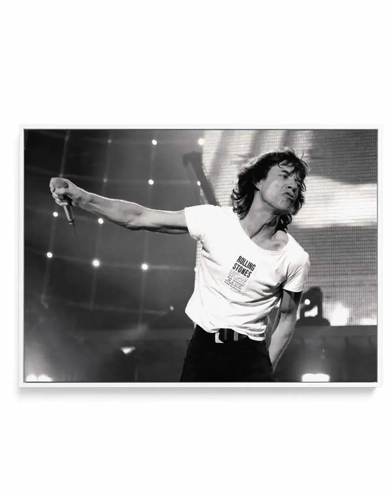 Framed black and white canvas print of Mick Jagger performing on stage, captured mid-song with a microphone in his outstretched left hand. He wears a white t-shirt with "ROLLING STONES" printed on it and black trousers, his long hair slightly disheveled. The background features bright stage lights and a large screen, creating a dynamic concert atmosphere. The print is displayed in a thin white frame.