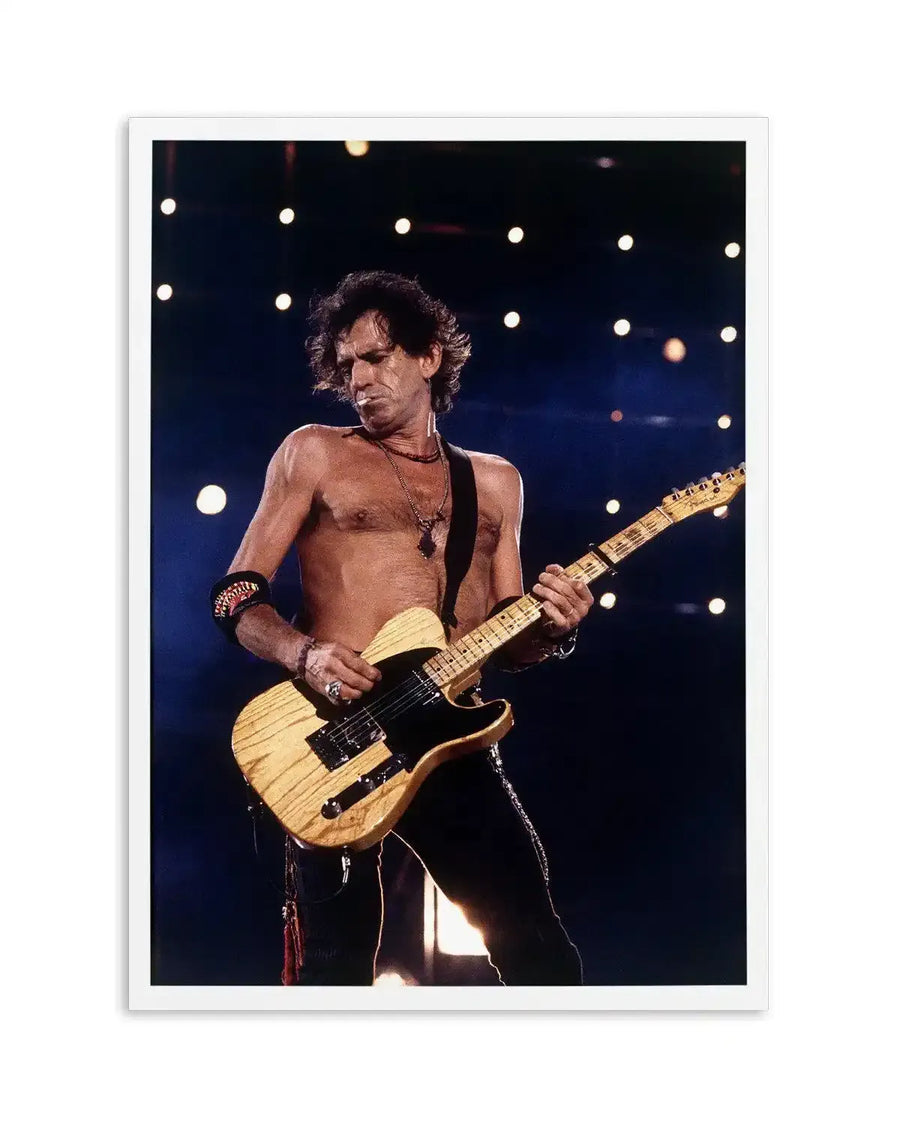 "Colour photograph art print of Keith Richards performing shirtless on stage, playing a blonde Fender Telecaster guitar. He is looking down at his guitar, with a cigarette hanging from his lips. He wears a dark necklace with a pendant and a dark armband on his left arm. The stage lights create a bokeh effect in the dark background. The print has a white border."