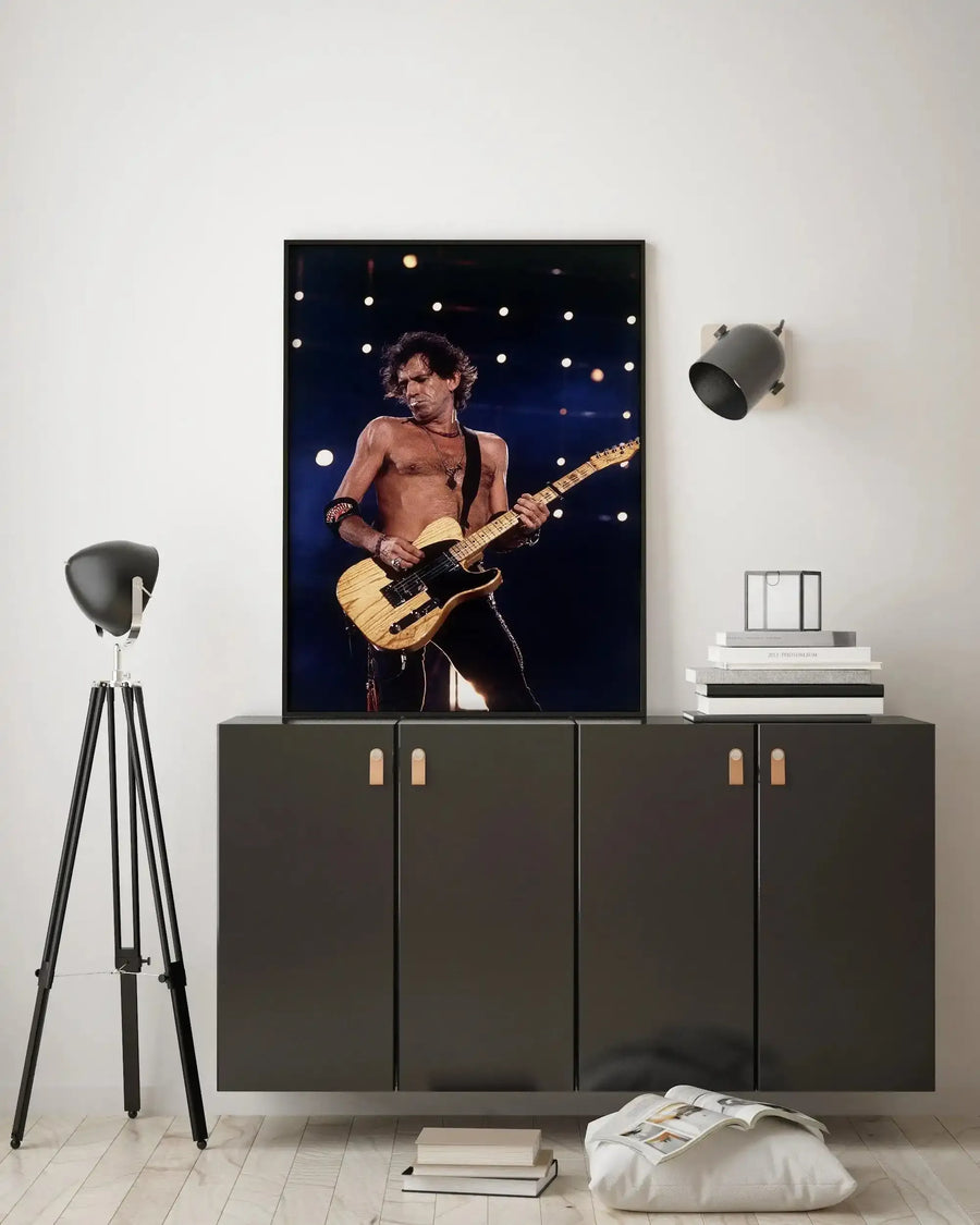 Lifestyle shot of a framed art print displayed above a dark grey cabinet in a minimalist room. The print features a photograph of Keith Richards playing guitar on stage, shirtless, with a light wood finish electric guitar against a dark background with blurred white and yellow stage lights. The room includes a black tripod floor lamp on the left and a black wall-mounted spotlight on the right, with a stack of books and a clear glass box on the cabinet.