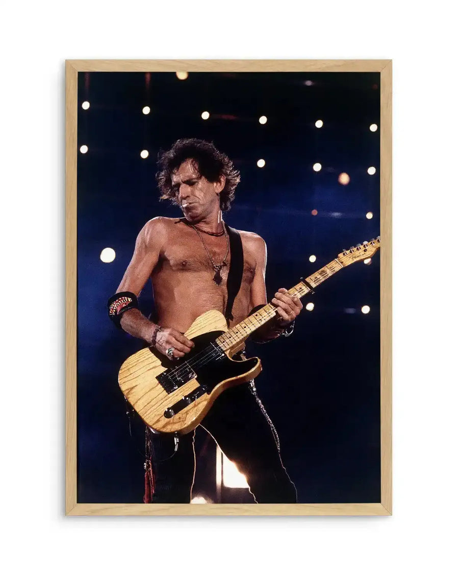 Framed photograph of musician Keith Richards playing an electric guitar on stage, displayed in a natural oak frame with white border. The composition features a shirtless Richards with dark, curly hair, wearing a necklace and armbands, looking down at his light wood-toned guitar with a black pickguard. The background is dark with bright, circular stage lights creating a dynamic, energetic mood.