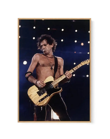 "Framed canvas print of a live concert photograph of Keith Richards of The Rolling Stones, playing an electric guitar under stage lights. Richards is shirtless, wearing dark pants and a necklace with a cross pendant, with a serious expression as he plays a light wood-toned electric guitar. The background is dark with bright white circular stage lights. The print is displayed in a natural wood frame."