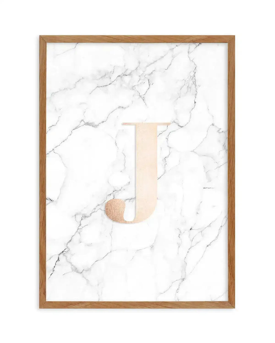 Framed art print featuring the letter "J" on a white marble background with gray veining. The letter is composed of a cream-colored vertical rectangle topped with a rose gold circular element. Wood-toned frame with white border. Portrait orientation.