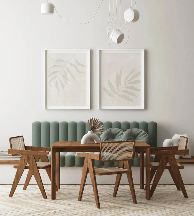 Lifestyle shot of a modern dining area featuring two framed Sage Botanica I art prints displayed above a sage green fluted bench seat. The prints feature a single large-scale botanical leaf in a muted sage green with subtle white outline against a warm cream background, with white frames and white borders. A wooden dining table with four wooden chairs is positioned in front of the bench, with a white arched vase holding dried palm fronds on the table.