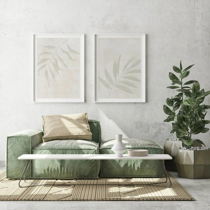 Lifestyle shot of a minimalist living room with two framed art prints displayed above a sage green sofa. The prints feature abstract botanical illustrations of single leaves in a pale sage green against a textured cream background, with white frames and white borders. The room includes a textured beige rug, a white coffee table with a white vase and a small bowl, and a large potted plant with dark green leaves in a geometric grey planter.