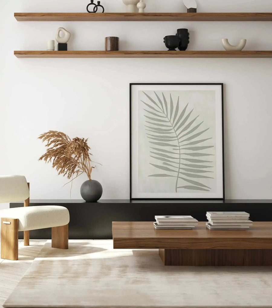 Lifestyle shot of a minimalist living room featuring a large framed art print of a sage leaf. The print depicts a single, large palm-like leaf with multiple fronds in a muted sage green against a pale green-grey background, displayed in a black frame with a white border. The print rests on a low black console table, next to a cream upholstered chair with wooden legs and a dark grey vase holding dried pampas grass. A light wood coffee table with stacks of books sits on a light-coloured rug in the foreground.