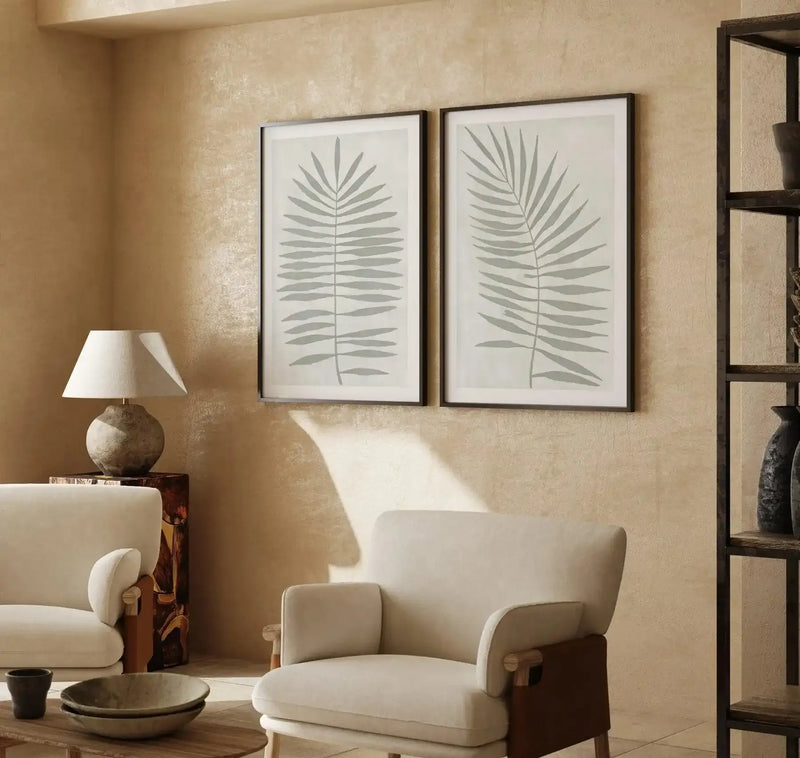Lifestyle shot of a minimalist living room with two framed art prints displayed on a textured beige wall. Each print features a single sage green palm leaf illustration with a black frame and white border. The room includes a cream armchair, a table lamp with a cream shade, and a dark wooden side table.