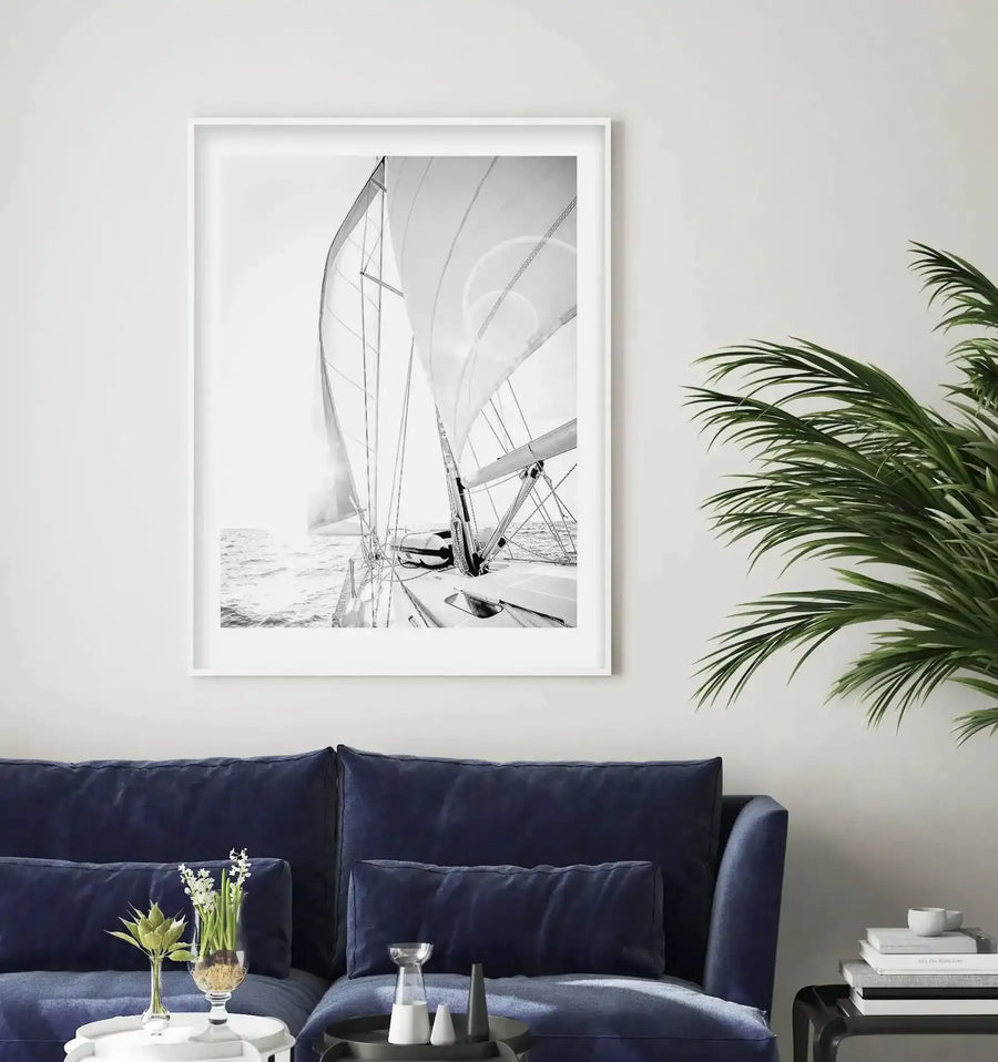 Lifestyle shot of a minimalist living room featuring a large framed black and white photograph of a sailboat on the ocean, displayed above a deep navy blue sofa. The print captures the bright sun reflecting off the water, with the boat's sails and rigging prominent in the foreground. A green palm plant is visible on the right, and a white side table with decorative items is on the left.