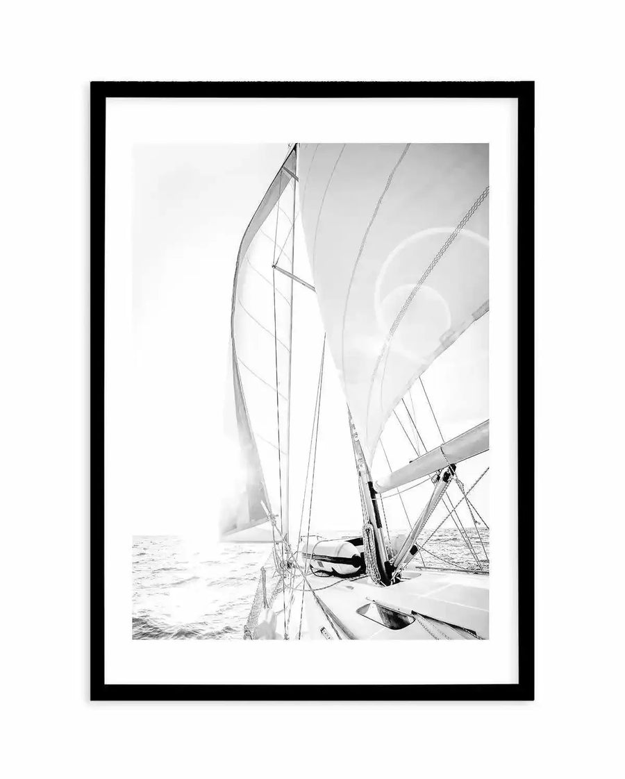 Framed black and white photograph of a sailboat on the open ocean with a bright sun flare. The composition shows the deck and sails of the boat from a low angle, with the sun low on the horizon creating a strong glare on the water and through the sails. The image is framed in black with a white border.