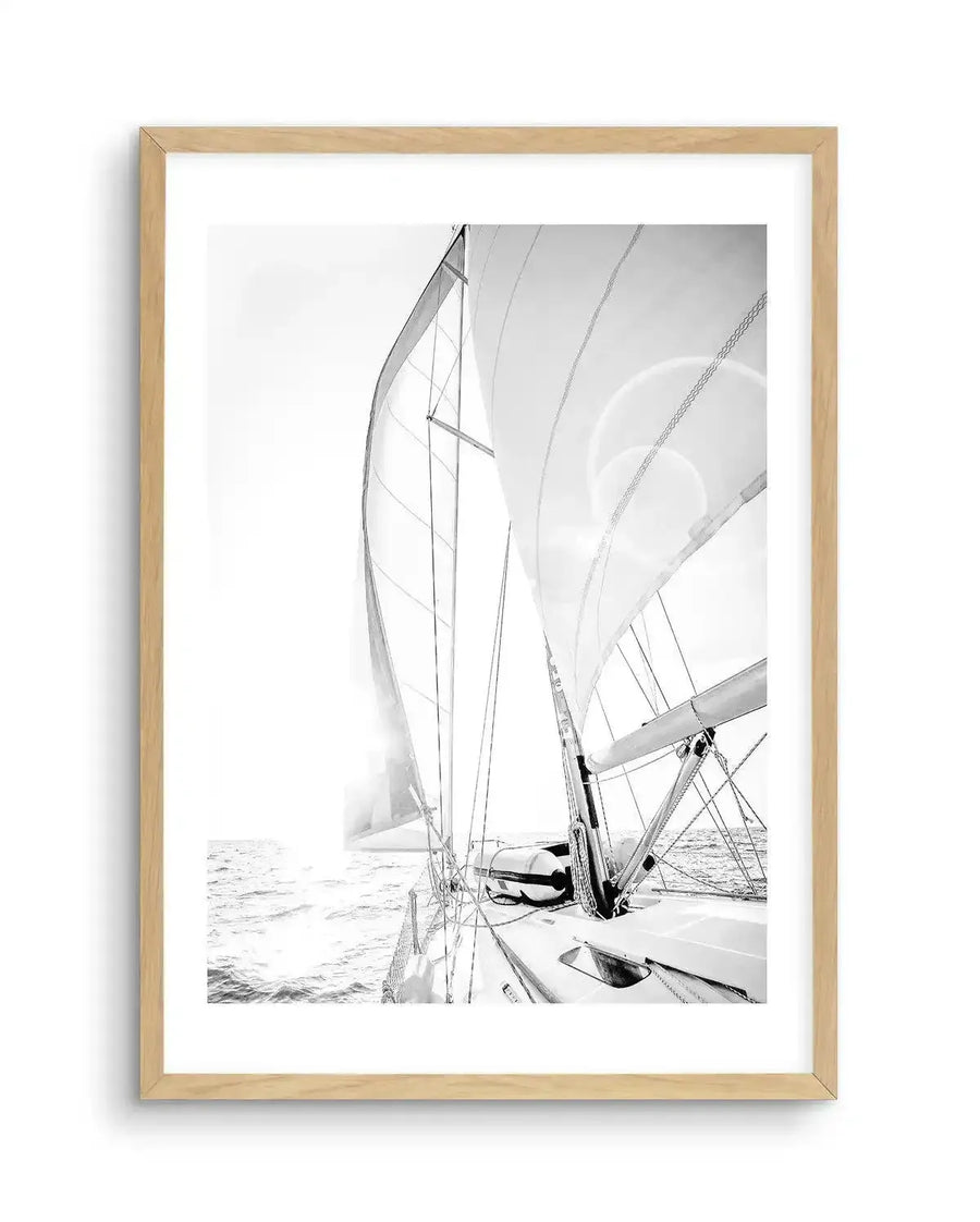 Framed black and white photograph of a sailboat on the open ocean with natural wood frame and white border. The composition captures the perspective from the deck of the boat, looking forward towards the sails, with the sun creating a bright lens flare on the right sail. The ocean is visible on the left, with gentle waves reflecting the bright light.