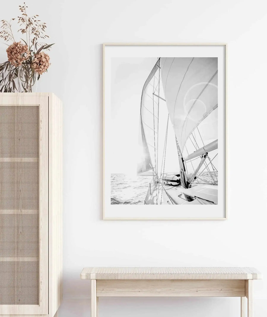 Lifestyle shot of a minimalist room with a large framed art print displayed above a light wood bench. The print is a black and white photograph of a sailboat on the ocean with sails up, viewed from the deck, with the sun creating a bright flare on the left. The print is in a natural wood frame with a white border. To the left, a light wood cabinet with a woven cane door is visible, topped with a glass vase holding dried flowers and grasses.