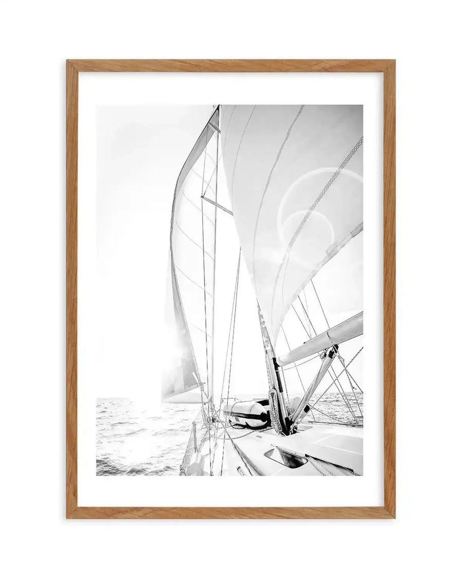 Framed black and white photograph of a sailboat on the open ocean with natural wood frame and white border. The composition is taken from the deck of the boat, looking forward and slightly to the left, with the large main sail and jib sail billowing in the wind. The sun is low in the sky, creating a bright glare on the water and through the sails.