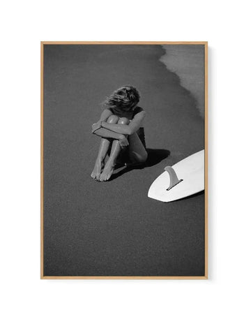 Black and white framed canvas art print titled "Salty" by Mario Stefanelli, featuring a woman sitting on dark sand with knees drawn up and arms wrapped around her legs, her head turned away from view. A white surfboard is positioned in the lower right corner. The photograph has high contrast with deep charcoal tones and bright white highlights. The canvas is mounted in a natural wood frame with a vertical orientation.
