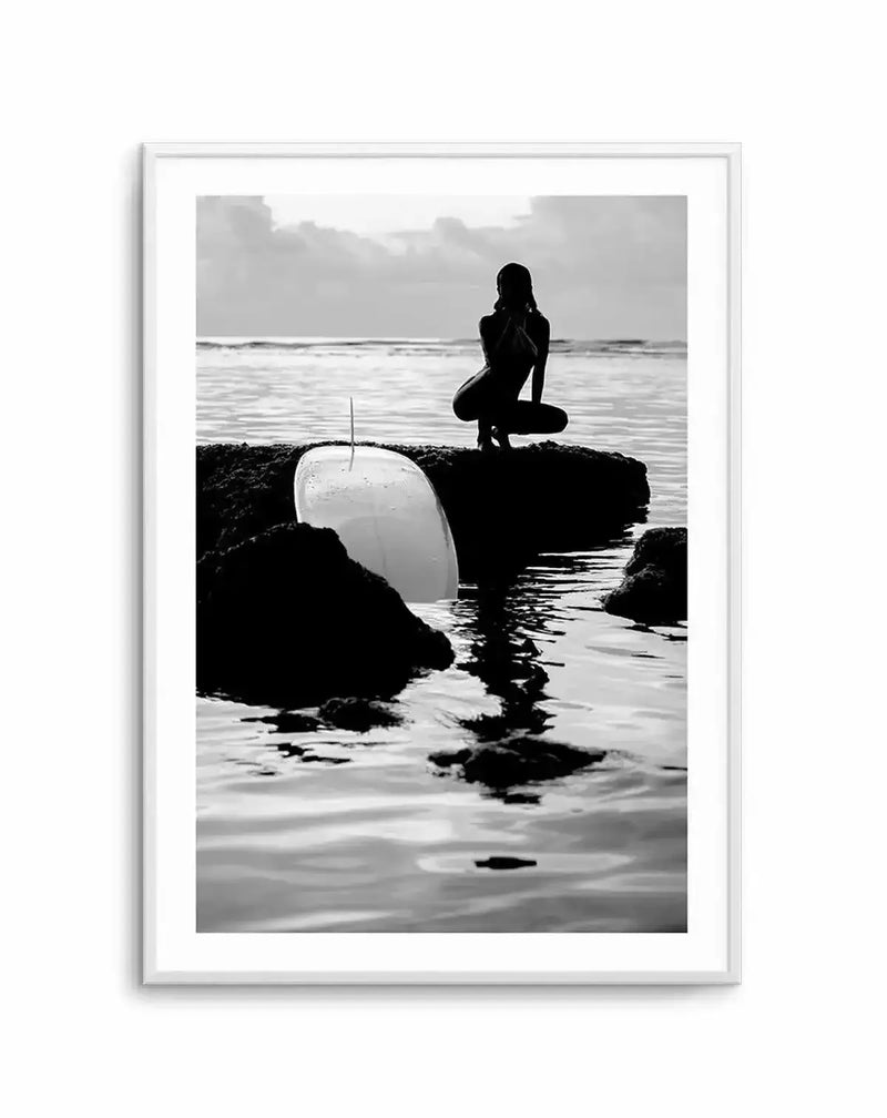 Black and white photograph titled "Salty II" by Mario Stefanelli, displayed in a silver frame with white border. The image shows a silhouetted female figure in a wetsuit crouching on dark rocky outcrops in shallow ocean water, holding a white surfboard. Overcast sky and calm sea create a moody, minimalist composition with strong contrast between the dark rocks and figure against the lighter water and cloudy backdrop.