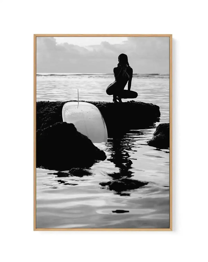 Black and white framed canvas art print titled "Salty II" by Mario Stefanelli, featuring a silhouetted female surfer in a wetsuit crouching on dark rocky outcroppings along a beach. A white surfboard leans against the rocks in the foreground. Calm ocean waters and overcast sky fill the background. The photograph has high contrast with deep blacks and bright whites, creating a moody, contemplative coastal scene. Natural wood-toned frame with white border surrounding the image.