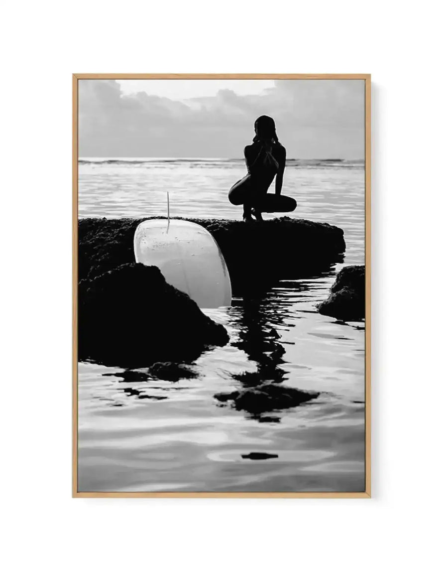 Black and white framed canvas art print titled "Salty II" by Mario Stefanelli, featuring a silhouetted female surfer in a wetsuit crouching on dark rocky outcroppings along a beach. A white surfboard leans against the rocks in the foreground. Calm ocean waters and overcast sky fill the background. The photograph has high contrast with deep blacks and bright whites, creating a moody, contemplative coastal scene. Natural wood-toned frame with white border surrounding the image.