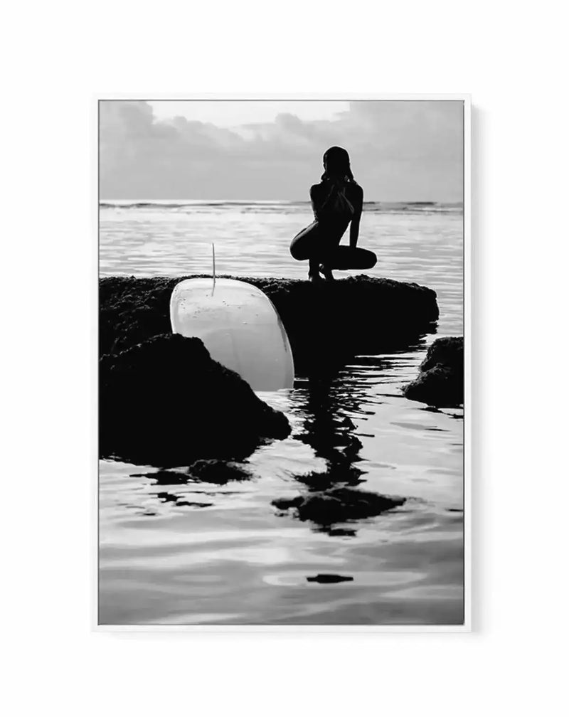 Black and white photograph titled "Salty II" by Mario Stefanelli, printed on canvas with gray frame and white border. The composition features a silhouetted female figure in a wetsuit crouching on dark rocky outcrops in shallow ocean water, holding a white surfboard. Calm waves and cloudy sky create a moody, contemplative seascape in the background. The high-contrast monochromatic aesthetic emphasizes the interplay of light on water and textured rock formations.