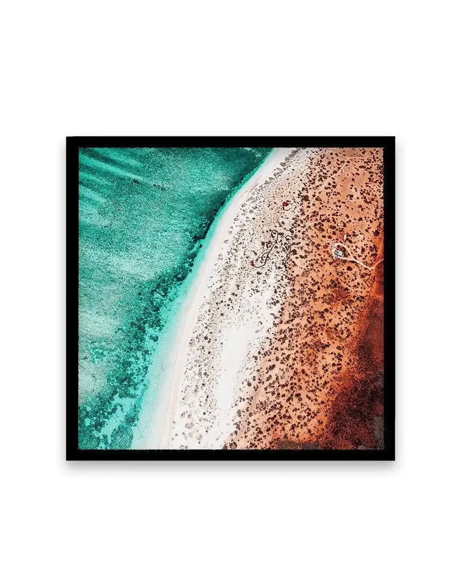 Aerial photograph art print in black frame showing a turquoise ocean transitioning to a white sand strip and reddish-brown desert landscape. The composition displays deep teal water with white foam meeting pale sand, which grades into rust and burnt orange tones of arid terrain. Captured from above, the image creates diagonal bands of color with rich texture and natural contrast. Square format with black border.