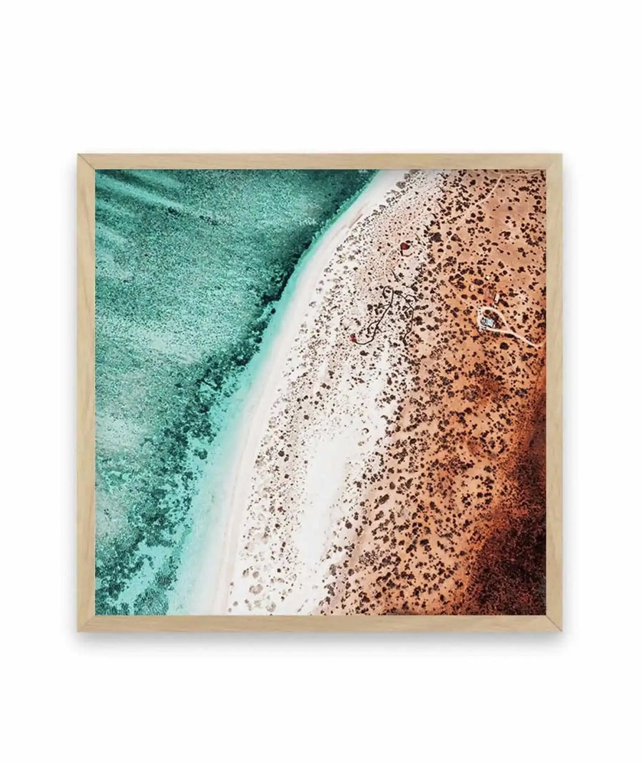 Aerial photograph framed in natural wood showing a curved white sandy beach shoreline dividing turquoise shallow water on the left from rust-brown dry sand dotted with seaweed on the right. The image captures the stark contrast between the wet and dry zones of the beach, with a small white figure visible on the sand. Square format with natural wood frame.
