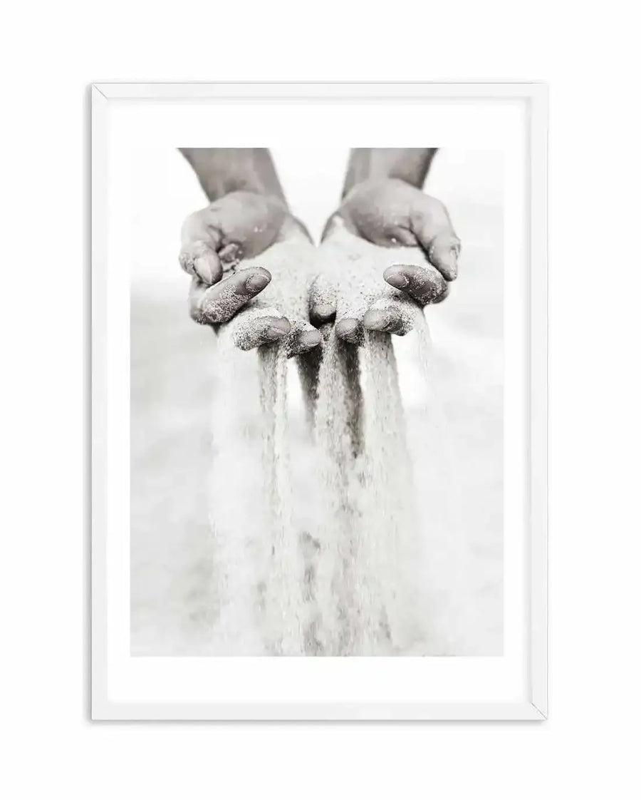 Framed black and white photograph of two hands cupped together, letting fine white sand pour through the fingers. The hands are slightly blurred, suggesting motion, and the sand creates delicate streams against a bright, soft white background. The print is displayed in a white frame with a white border.