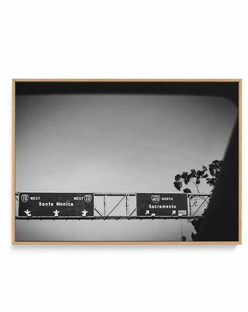 Black and white photograph of highway road signs mounted on an overpass against a clear sky, with a bare tree visible on the right. One sign reads "10 WEST Santa Monica" with directional arrows, the other reads "405 NORTH Sacramento ONLY" in white text on dark backgrounds. The image is mounted in a natural wood frame with a white border.