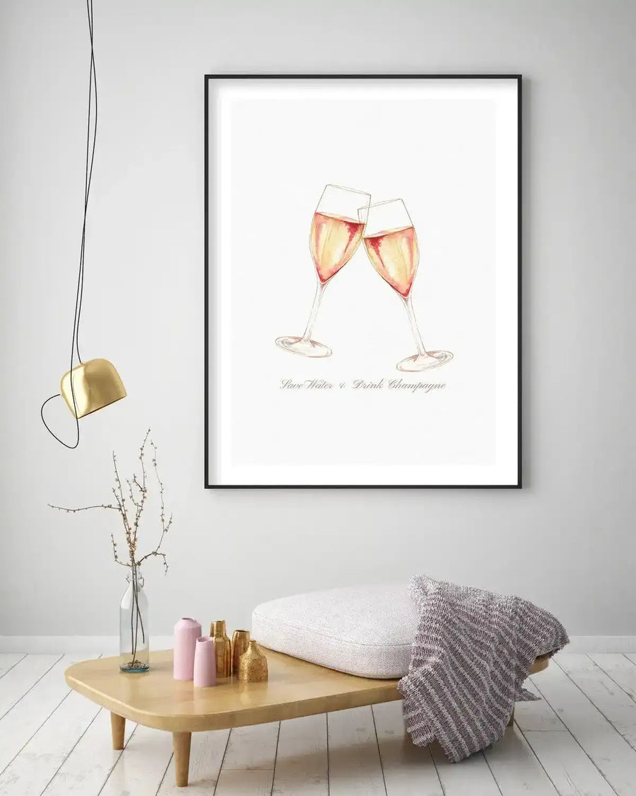 Lifestyle shot of a minimalist living room featuring a framed art print above a low wooden coffee table. The print depicts two watercolour champagne flutes clinking, filled with a pale rose-gold liquid, with the text "Save Water & Drink Champagne" in a delicate script below. The print is displayed in a black frame with a white border. The room includes a gold pendant light, a glass vase with dried branches, pink and gold decorative vases, a white cushioned bench, and a grey and white striped throw blanket.