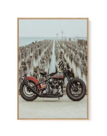 Framed canvas art print titled "Sayonara By Tim Harris" featuring a vintage red and black motorcycle with white-wall tires positioned in profile on a concrete surface. The motorcycle is centered in the composition against a blurred background of wooden pier pilings extending into calm water under an overcast sky. The image has a faded, vintage color palette with muted tones. The canvas is housed in a natural wood frame with a white border.