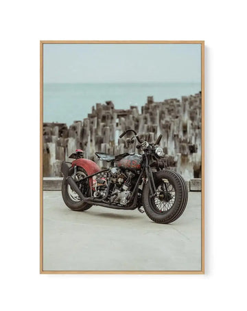 Framed canvas art print titled "Sayonara Pier By Tim Harris" featuring a vintage motorcycle with a red sidecar parked on a concrete pier. The motorcycle has dark weathered metal detailing and spoke wheels. Behind it stretches a dense urban cityscape with tall apartment buildings in cream and white tones meeting a pale turquoise sea and misty sky. The photograph has a faded, nostalgic color palette with soft muted tones. Natural wood frame with light tan finish surrounds the canvas.