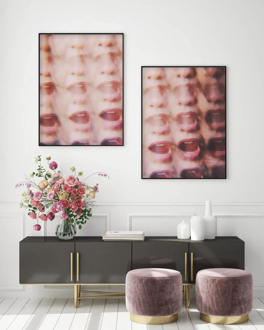 Two framed art prints displayed above a dark grey console table in a modern living room. The prints feature a repeating pattern of blurred female faces with open mouths and dark red lips, creating a distorted, abstract effect. The frames are black with no border. The room includes a large vase of pink and yellow roses, white decorative vases, and two round velvet ottomans in dusty rose with gold bases.